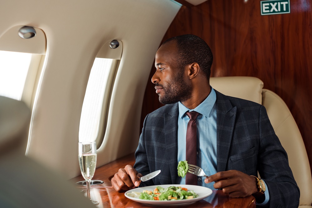 handsome african american businessman holding cutlery near salad in private jet Business man eating in business class and looking out the window