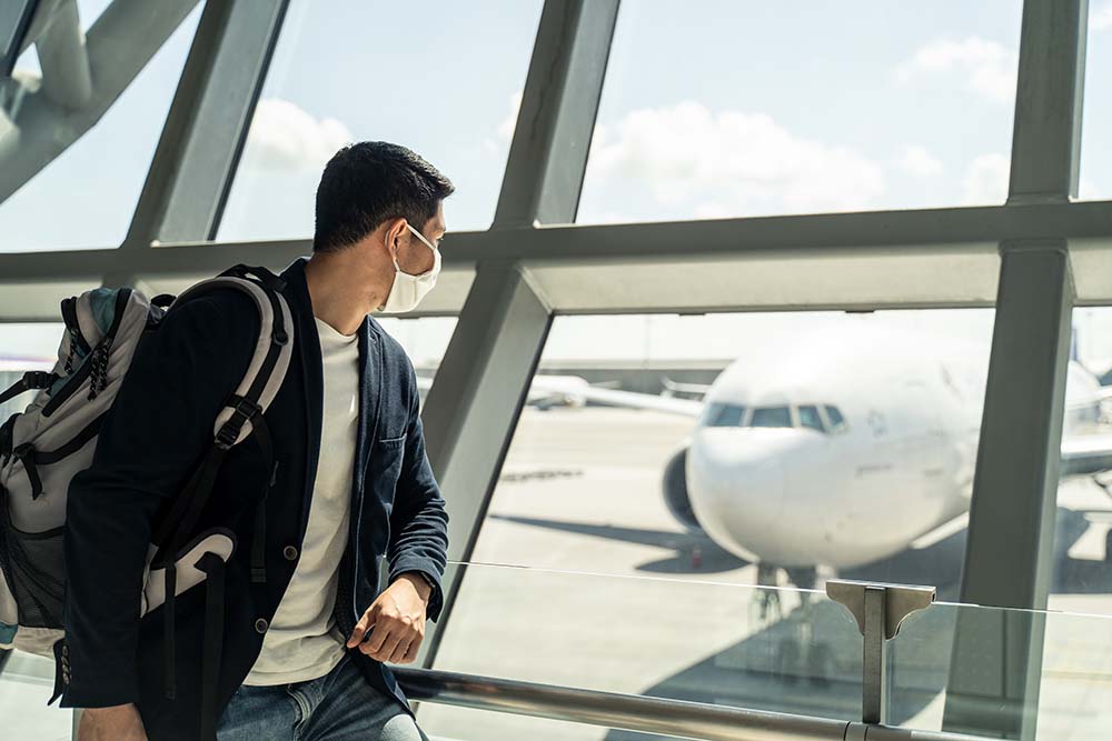 Asian traveler business man wearing face mask waiting to board i Man with mask on carrying a backpack looking at plane from inside an airport