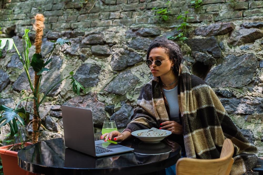 african american freelancer in sunglasses covered in blanket using laptop and having lunch in cafe Mädchen arbeitet remote aus einem Café.