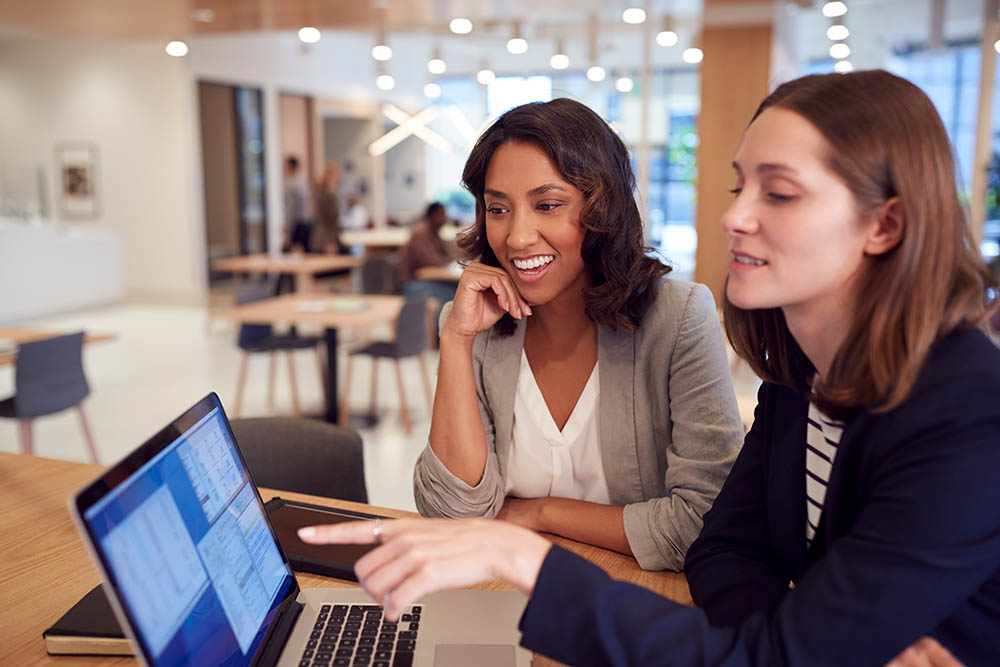 Two Businesswomen With Laptop At Desk In Open Plan Office Collaborating On Project Together two travel managers planning business trips