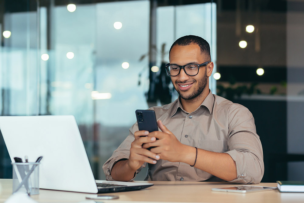 Happy african american businessman in glasses working inside modern office with laptop, man in shirt sitting at table browsing online pages using smartphone Man with glasses looking at cellphone in the office