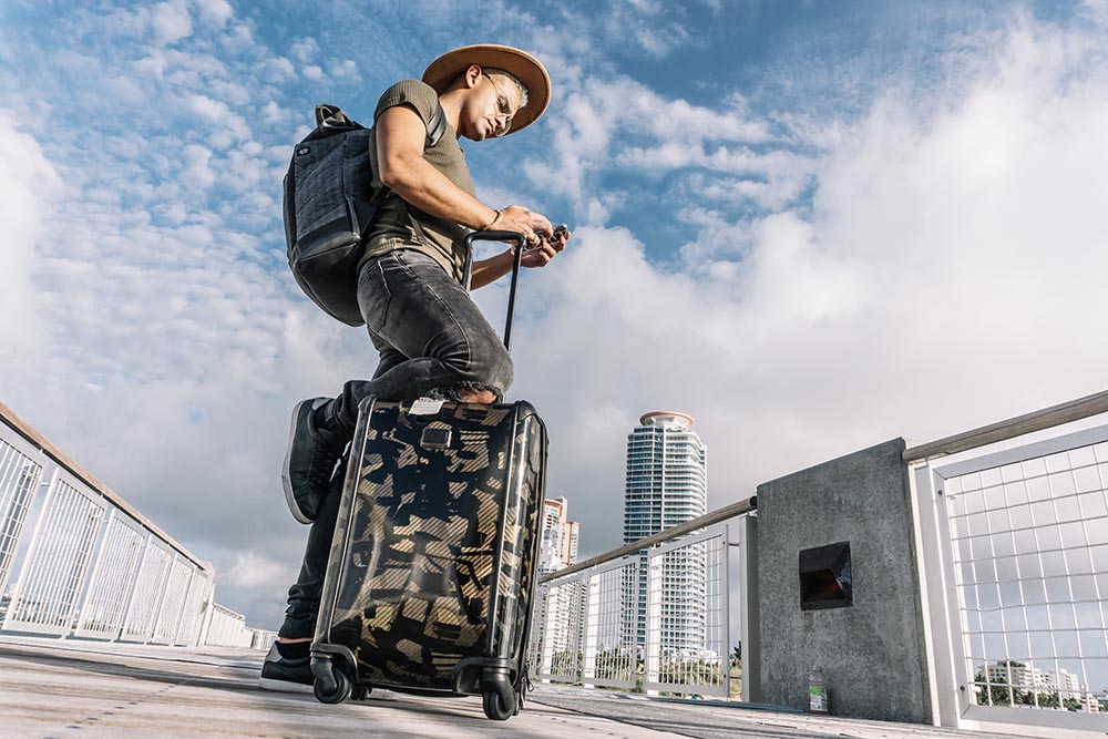 Traveller man texting phone with his luggage walking on Ocean drive South Beach Florida Geschäftsreisender mit Koffer trägt Hut und schaut auf sein Mobiltelefon
