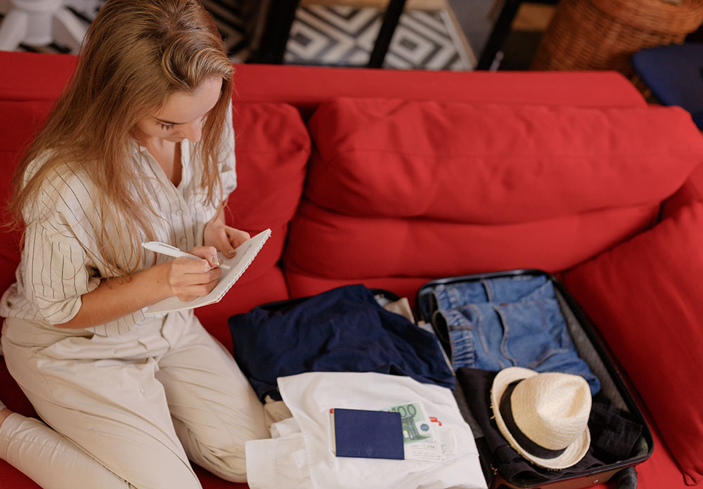 Young woman checking accessories and stuff in luggage on couch at home Junge Frau, die zu Hause auf der Couch Accessoires und Sachen im Gepäck überprüft
